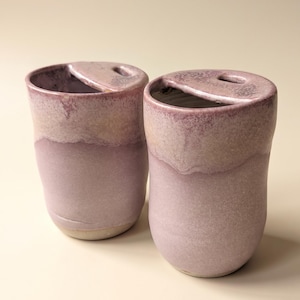 May include: Two ceramic tumblers with a soft, matte mauve glaze. Each vessel features a unique, integrated lid design with a small, oval-shaped opening at the top. The finish transitions from a deeper, textured purple at the rim to a lighter, muted tone.