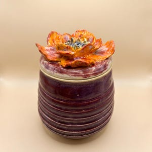 Puede incluir: Un tarro de cerámica con cuerpo acanalado de color morado intenso y tapa a juego. La tapa está rematada con una gran flor esculpida de color naranja y amarillo con un centro oscuro. El esmalte del tarro y la tapa tiene un acabado brillante.