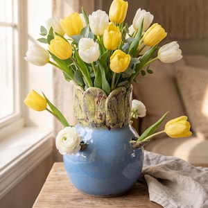 May include: A ceramic vase with a glossy blue base and a textured, leaf-like green rim. The vase features multiple openings for stems and is filled with a bouquet of yellow and white tulips, along with small white ranunculus flowers.