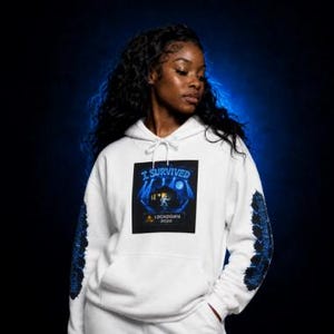 Op de afbeelding: Een wit sweatshirt met capuchon en een centrale afbeelding met de tekst I SURVIVED LOCKDOWN 2020, met een figuur in een donkerblauw landschap. De mouwen hebben donkerblauwe patronen die over de gehele lengte van elke arm lopen.
