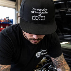 Peut inclure: Une casquette de baseball noire avec le texte brodé en blanc "You can blow my head gasket" au-dessus d'une illustration graphique blanche d'un joint de culasse de moteur à quatre cylindres avec une petite fumée au centre.