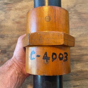 May include: A wooden object with a cylindrical shape, featuring a light brown body and black ends. The text "C-4903" is hand-painted on the side. The object is held in a person's hand.