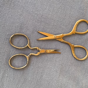 May include: Two pairs of gold-colored metal scissors with ornate, textured handles. One pair features straight, pointed blades, while the other pair has a distinct curved design with a small screw at the pivot point, showcasing a metallic finish.