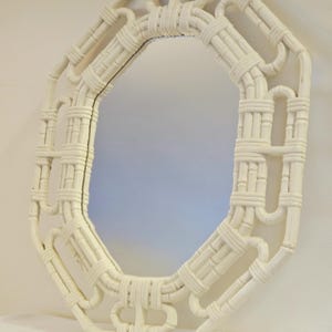 May include: An octagonal wall mirror featuring a white frame constructed from woven rattan-style material. The frame displays a geometric pattern with interconnected loops and straight segments, surrounding a central reflective glass surface.