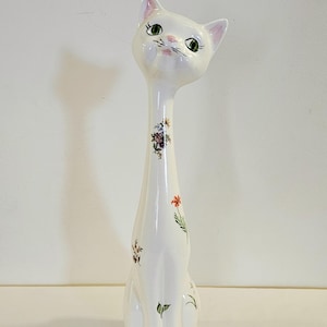May include: A tall, white ceramic cat figurine with an elongated neck, featuring painted green eyes, pink ears, and delicate floral patterns in various colors scattered across its body. The cat is in a seated position with a smooth, glossy finish.