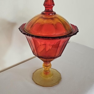 Könnte beinhalten: Eine Vintage-Bonbonniere mit Deckel in Rot- und Gelbtönen. Die Glasschale hat eine facettierte Schale, einen dekorativen Rand und einen konischen Knauf auf dem Deckel. Der Sockel ist hellgelb und die Schale wird von einem Stiel getragen.