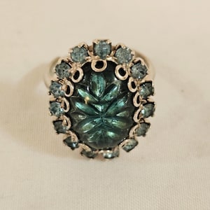 May include: A silver-toned ring featuring a large, oval-shaped, dark teal glass cabochon with a carved leaf-like pattern. The central stone is surrounded by a halo of small, round, light blue faceted crystals set in a scalloped metal frame.