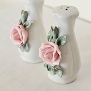 May include: A pair of white ceramic salt and pepper shakers, each featuring a three-dimensional sculpted pink rose with sage green leaves attached to the front. The shakers have a smooth, glossy finish and a slightly flared base and top.