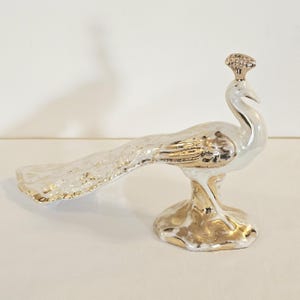 May include: A ceramic peacock figurine featuring a glossy white finish with metallic gold accents on the crest, neck, wings, tail feathers, and base. The bird is depicted in a standing position with its long tail extended horizontally to the left.