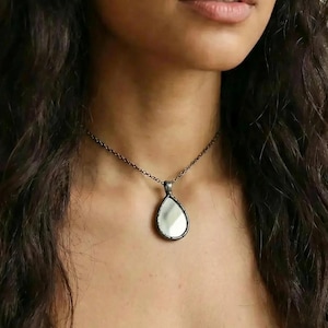 May include: A silver-coloured necklace featuring a teardrop pendant. The pendant has a mirrored surface. The woman in the image has long, dark hair and is looking directly at the camera.