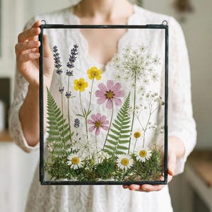 May include: A framed botanical art piece featuring pressed flowers and greenery. The arrangement includes lavender, yellow and pink flowers, and ferns, all set against a clear background within a black metal frame. The artwork is a square shape.