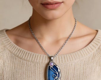 May include: A silver-toned pendant necklace featuring an elongated oval blue agate stone with dark blue banding. The stone is encased in a decorative silver metal frame with swirling wire accents and a small, round, iridescent purple cabochon at the top.
