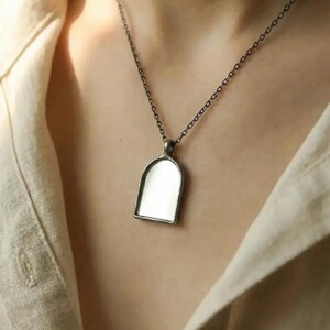 Op de afbeelding: Een zilverkleurige hangerketting met een rechthoekig frame en een boogvormige bovenkant. De hanger heeft een reflecterend, spiegelachtig oppervlak. De ketting wordt gedragen over een neutraal gekleurd overhemd.