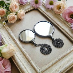 May include: Two round mirrors with dark metal frames and attached dark metal chains. The mirrors are of different sizes, each featuring a smooth reflective surface and a soldered metal border, presented within a weathered, light-colored wooden frame.