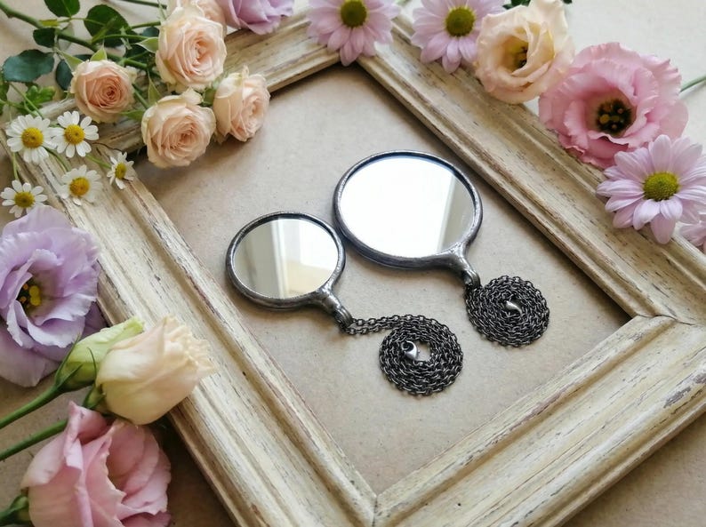 May include: Two round mirrors with dark metal frames and attached dark metal chains. The mirrors are of different sizes, each featuring a smooth reflective surface and a soldered metal border, presented within a weathered, light-colored wooden frame.