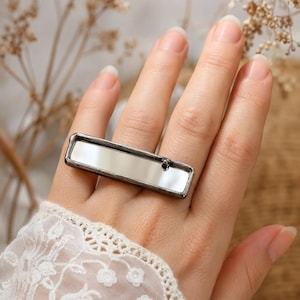 Op de afbeelding: Een rechthoekige ring met een reflecterend spiegeloppervlak in een donker, geoxideerd metalen frame. Het langwerpige, horizontale ontwerp ligt over de vinger en toont een gladde, gepolijste afwerking binnen de metalen rand.