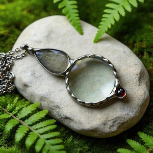 May include: A silver-toned magnifying glass pendant with a clear lens and a small red gemstone. The pendant is on a silver chain and rests on a light gray stone surrounded by green moss and fern leaves. A unique piece of jewelry.