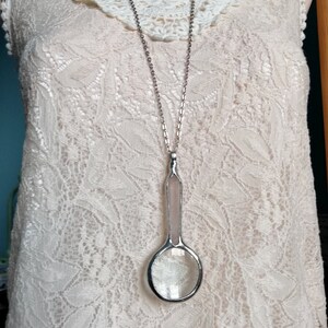 May include: A long silver necklace with a magnifying glass pendant. The pendant has a clear glass lens and a light pink handle. The necklace is displayed on a cream-colored lace dress with a crochet collar.