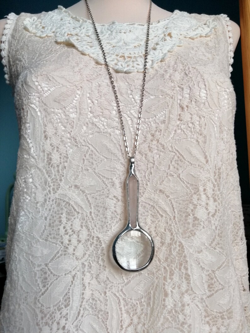May include: A long silver necklace with a magnifying glass pendant. The pendant has a clear glass lens and a light pink handle. The necklace is displayed on a cream-colored lace dress with a crochet collar.