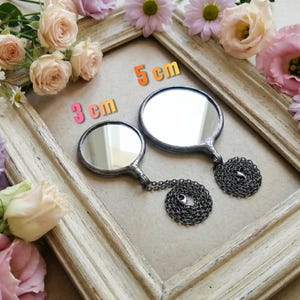 May include: Two round mirrors with dark metal frames and attached dark metal chains. One mirror is labeled 3 cm and the other is labeled 5 cm. The mirrors are positioned within a light-colored wooden frame surrounded by pink and white flowers.