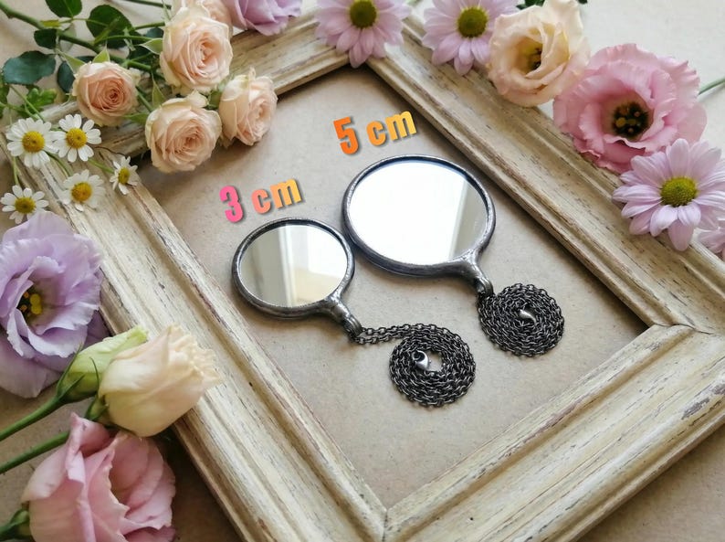 May include: Two round mirrors with dark metal frames and attached dark metal chains. One mirror is labeled 3 cm and the other is labeled 5 cm. The mirrors are positioned within a light-colored wooden frame surrounded by pink and white flowers.