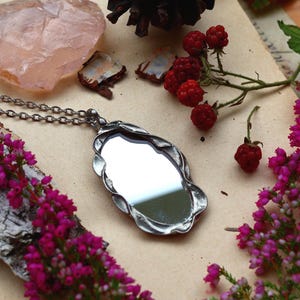 Peut inclure: Un collier pendentif en métal argenté doté d'un miroir de forme ovale serti dans un cadre texturé à l'aspect organique. Le pendentif est suspendu à une chaîne assortie en métal argenté, révélant une surface réfléchissante.