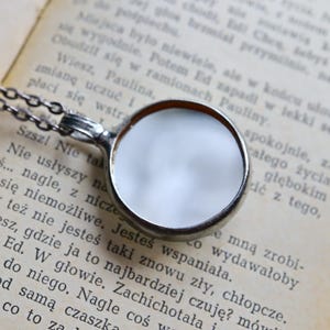 May include: A silver-toned circular pendant necklace featuring a smooth, reflective metallic surface. The pendant is attached to a delicate silver-toned chain, creating a simple and clean design that rests against a page of printed text.