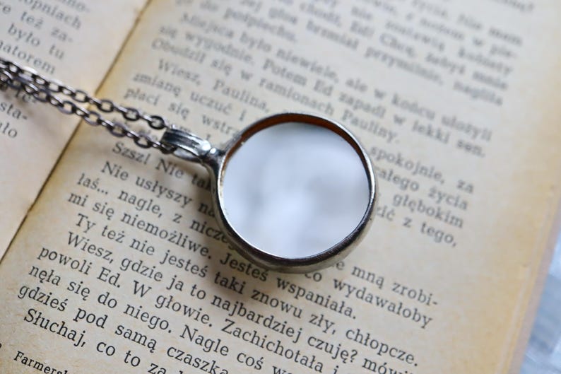 May include: A silver-toned circular pendant necklace featuring a smooth, reflective metallic surface. The pendant is attached to a delicate silver-toned chain, creating a simple and clean design that rests against a page of printed text.
