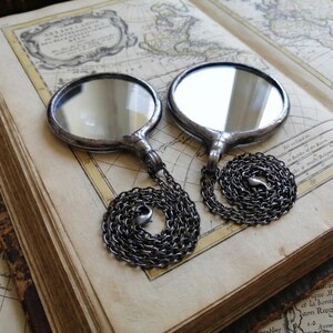 May include: Two circular, silver-toned metal framed mirrors, each featuring a reflective glass surface and a dark, coiled metal chain attached to the base. The mirrors are positioned side-by-side, showcasing a smooth, metallic finish and a simple, functional design with a clear, polished appearance.