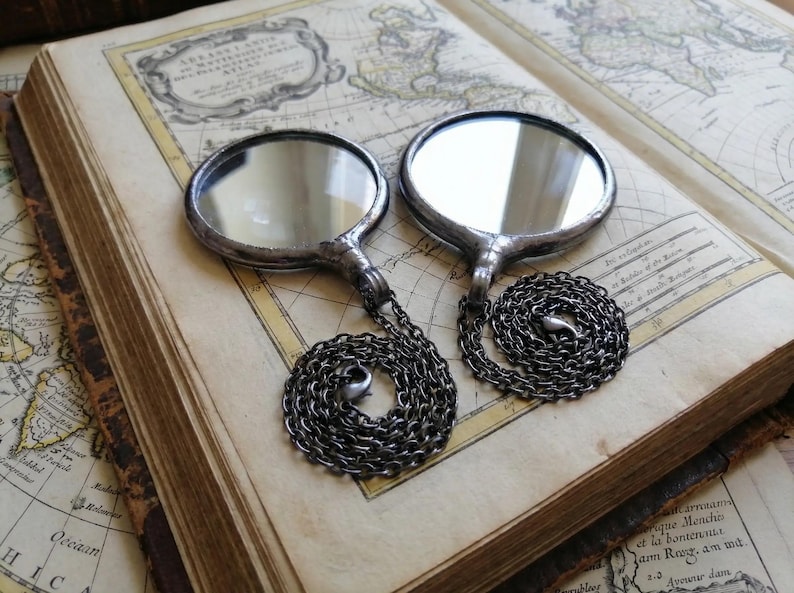May include: Two circular, silver-toned metal framed mirrors, each featuring a reflective glass surface and a dark, coiled metal chain attached to the base. The mirrors are positioned side-by-side, showcasing a smooth, metallic finish and a simple, functional design with a clear, polished appearance.