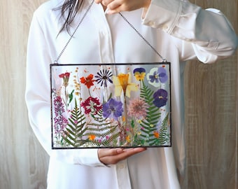 May include: A rectangular glass wall hanging featuring a collection of pressed dried flowers and green ferns encased between two clear glass panes with a dark metal frame and a thin black metal chain for suspension from the top corners.
