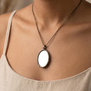 Peut inclure: Un collier pendentif de forme ovale doté d'une surface miroir réfléchissante encastrée dans un cadre en métal de couleur bronze foncé patiné. Le pendentif est suspendu à une chaîne en métal assortie avec de petits maillons ovales.