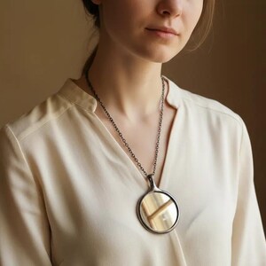 May include: A circular pendant necklace featuring a polished, reflective metallic disc encased in a dark, oxidized silver-toned frame, suspended from a matching dark metal chain that rests against a light-colored, long-sleeved blouse.