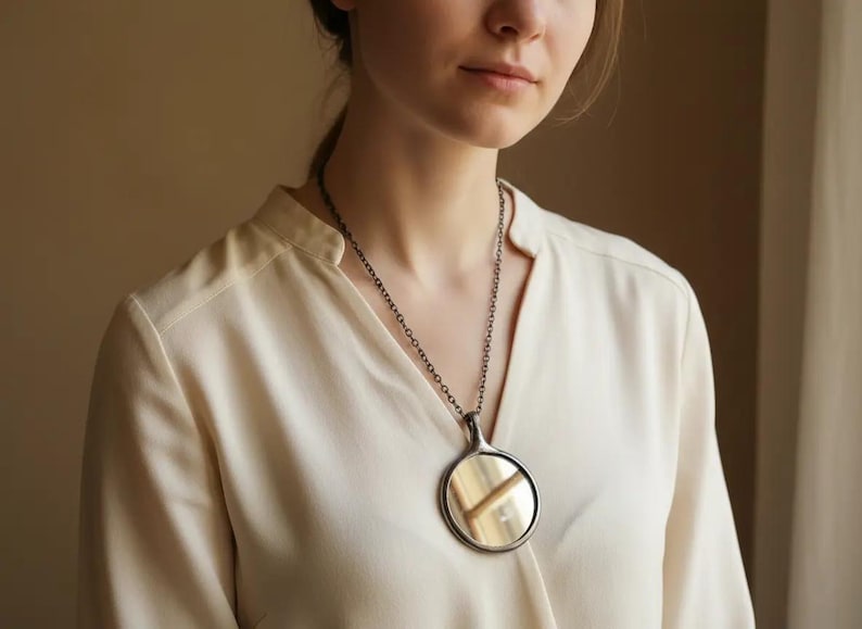 May include: A circular pendant necklace featuring a polished, reflective metallic disc encased in a dark, oxidized silver-toned frame, suspended from a matching dark metal chain that rests against a light-colored, long-sleeved blouse.