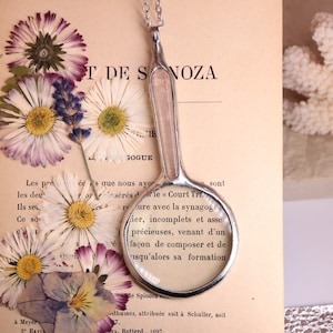 May include: A vintage-style magnifying glass with a silver handle and a clear lens, resting on an open book with dried flowers. The book's pages display French text. A white coral piece is in the background.
