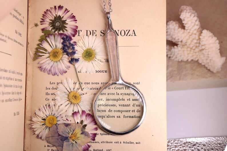 May include: A vintage-style magnifying glass with a silver handle and a clear lens, resting on an open book with dried flowers. The book's pages display French text. A white coral piece is in the background.