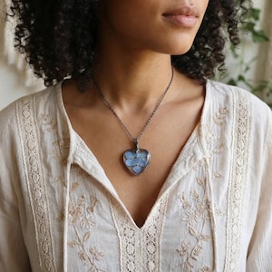 Peut inclure: Un collier argenté avec un pendentif en forme de cœur rempli de fleurs bleu clair. Le pendentif est suspendu à une fine chaîne. La personne porte un chemisier crème avec des détails floraux brodés et une bordure en dentelle.