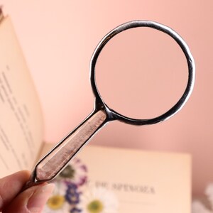 May include: A silver-toned magnifying glass with a rectangular handle and a circular lens. The handle has a light brown, patterned insert. The magnifying glass is held over an open book with text on the pages. The background is a soft pink.