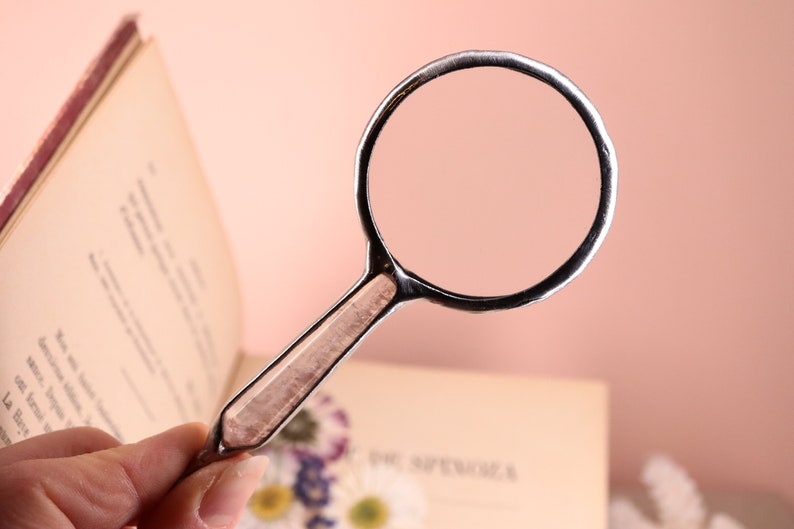 May include: A silver-toned magnifying glass with a rectangular handle and a circular lens. The handle has a light brown, patterned insert. The magnifying glass is held over an open book with text on the pages. The background is a soft pink.