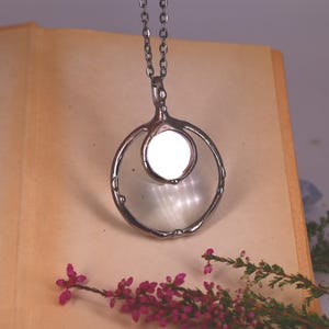 May include: A silver-toned pendant necklace featuring a large circular frame with a smaller, solid circular disc suspended within the center. The pendant hangs from a matching silver-toned chain, showcasing a metallic finish and a clean, circular design.