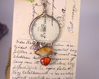 May include: A silver pendant necklace with a magnifying glass design. The pendant features a clear glass lens and is adorned with three colorful gemstones: orange, yellow, and a smaller iridescent stone. The background is a vintage postcard.