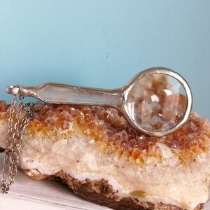 May include: A silver-toned magnifying glass with a round lens and a long handle rests on a rough, orange-yellow crystal formation. A delicate chain hangs from the handle. The background is a light blue.