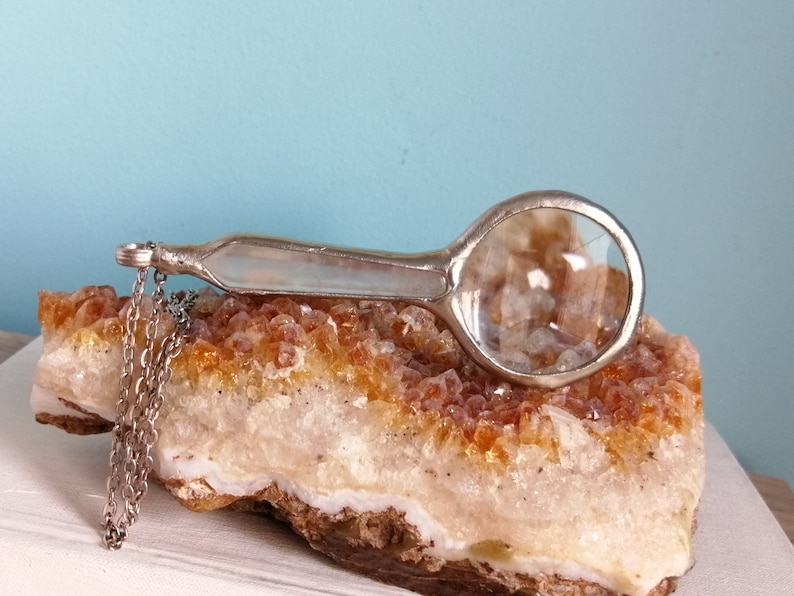 May include: A silver-toned magnifying glass with a round lens and a long handle rests on a rough, orange-yellow crystal formation. A delicate chain hangs from the handle. The background is a light blue.