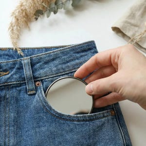 May include: A close-up of blue denim jeans with a small pocket. A hand is holding a small, round, silver-colored compact mirror, partially inserted into the pocket. Dried plants and a linen cloth are in the background.