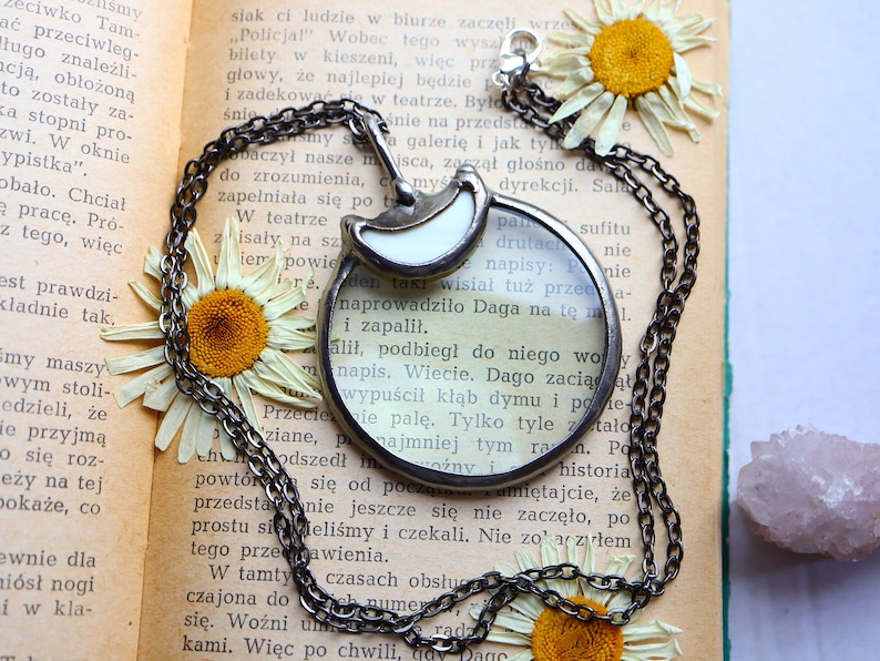 May include: A silver-toned magnifying glass pendant necklace with a crescent moon design, resting on an open book with dried daisies. The chain is dark silver. The pendant is approximately 2 inches in diameter.