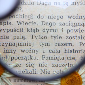 May include: Close-up of a page from a book with Polish text, magnified by a round magnifying glass. The page is aged with visible text and a daisy flower at the bottom. The text is in black ink.
