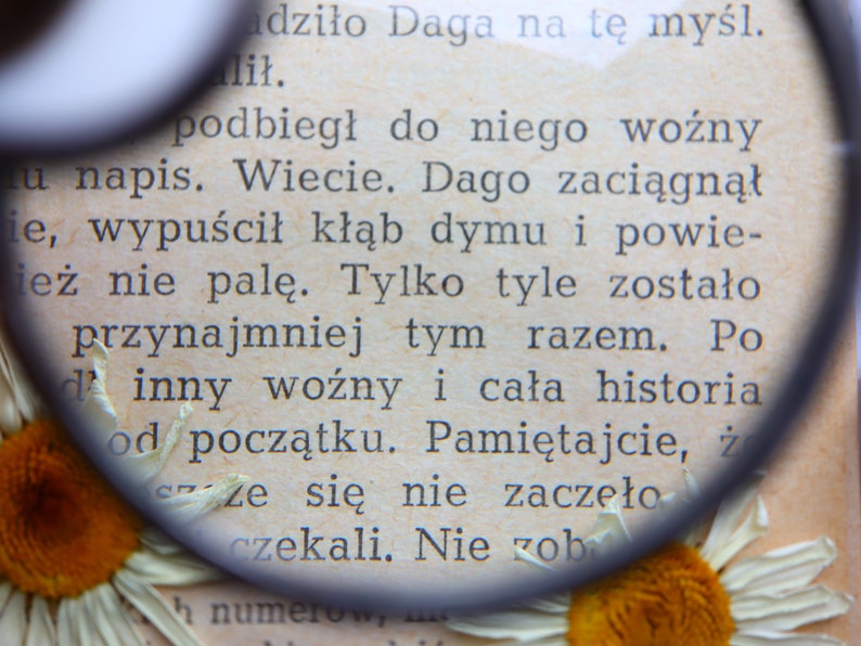 May include: Close-up of a page from a book with Polish text, magnified by a round magnifying glass. The page is aged with visible text and a daisy flower at the bottom. The text is in black ink.