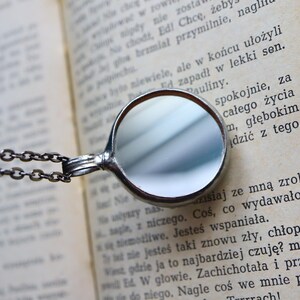 May include: A circular silver-toned pendant with a reflective mirror surface, attached to a dark metal chain. The pendant rests against an open book with printed text in a Latin-based language, showing clear black letters on aged off-white paper.
