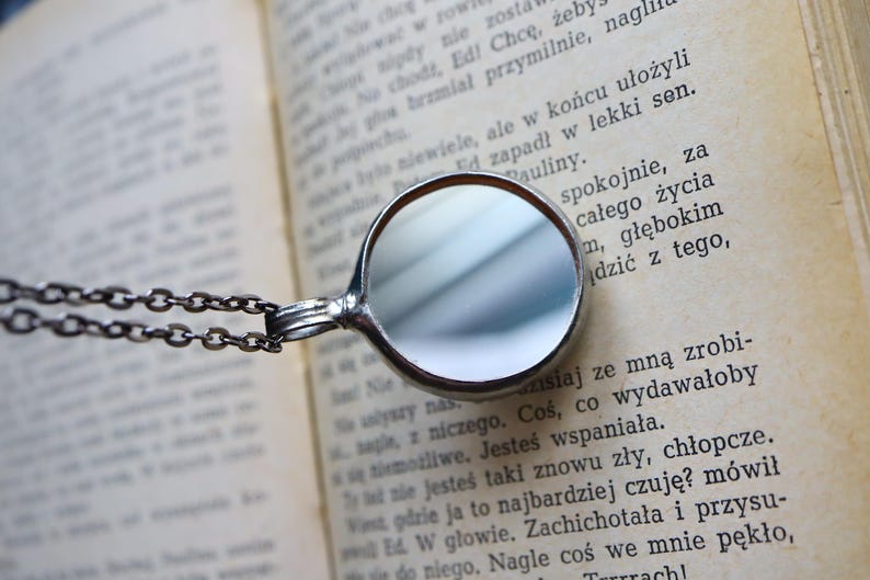 May include: A circular silver-toned pendant with a reflective mirror surface, attached to a dark metal chain. The pendant rests against an open book with printed text in a Latin-based language, showing clear black letters on aged off-white paper.