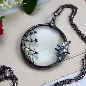 May include: A silver-toned magnifying glass pendant necklace. The round magnifying glass is set in a decorative frame with a chain. The pendant has a unique, artistic design, with a spiderweb-like pattern on the frame.
